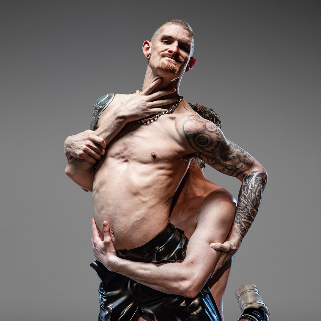 Two performers embrace in a tender reclining position. The male in the foreground has tattoos on each arm.